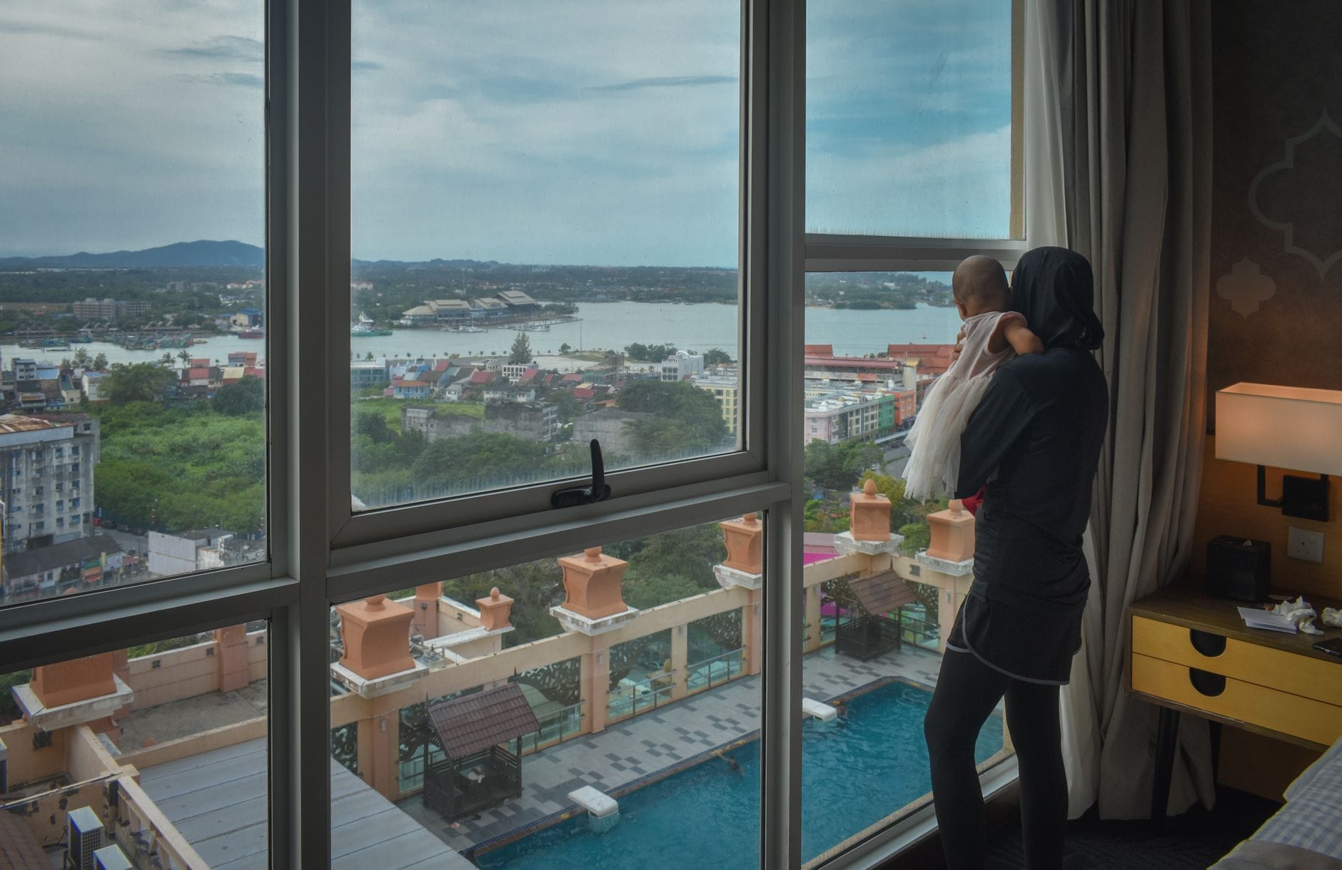 Mama z niemowlęciem patrzy z okna hotelu na miasto nad wodą